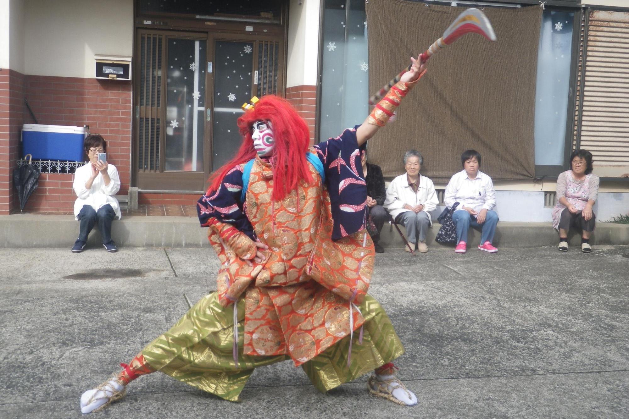 鎌を持ち、赤いかつらと独自の衣装を着て踊りを踊る人の写真